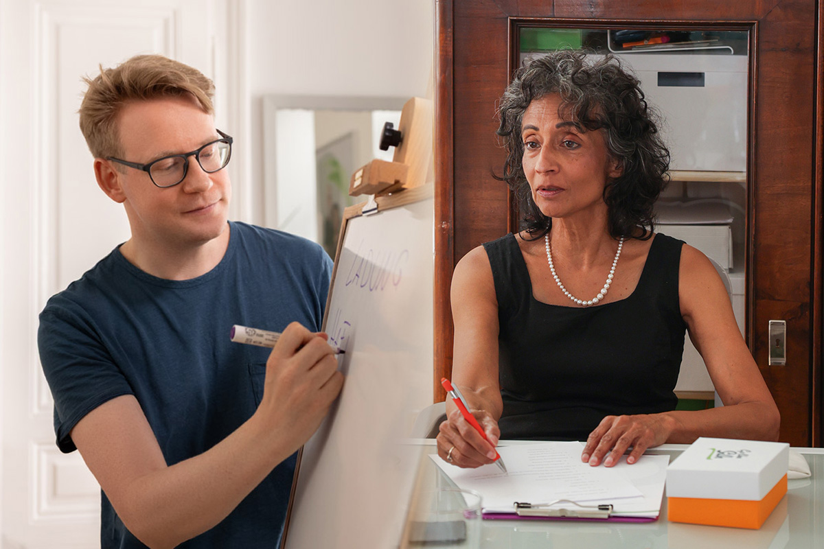 Michael En schreibt auf einem Whiteboard; Michèle Cooke sitz an einem Tisch und hält einen Stift in ihrer Hand.